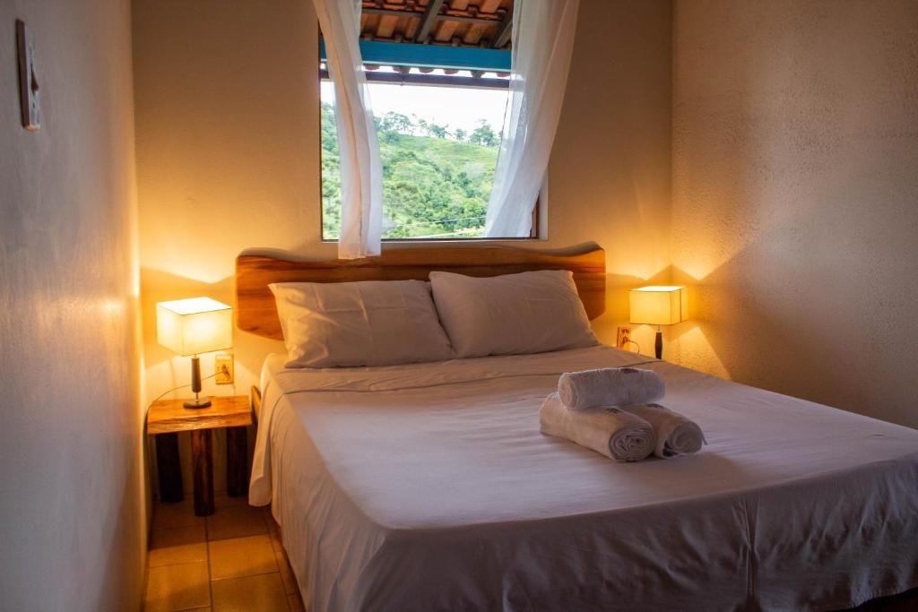 Suíte com cama de casal, abajures acesos e janela para o verde